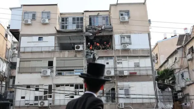 多層階の建物の壁が崩落し、その向こうでヘルメットをかぶった男性たちが作業している。その様子を、ユダヤ教超正統派の黒い帽子をかぶりと上着を着て、もみあげを伸ばした男性が地上から見上げている。