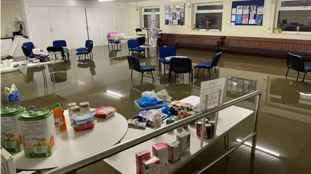 El interior del banco de alimentos después de la inundación. Sillas y mesas están sumergidas en el agua. Mesas largas con comida encima.