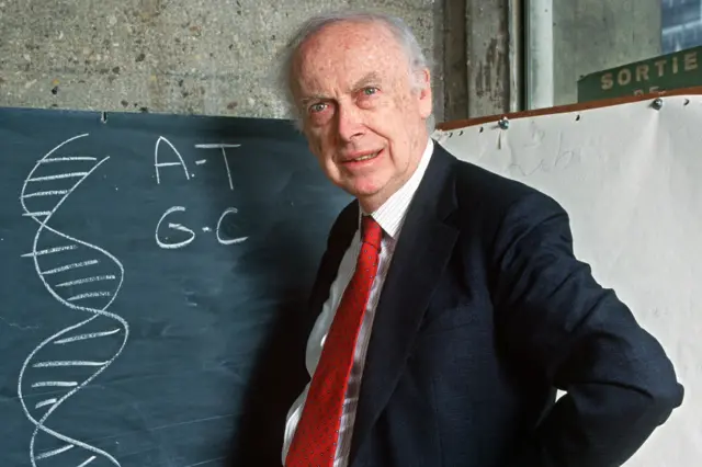 James Watson is wearing a dark suit jacket, white shirt, and red tie is standing beside a blackboard. The blackboard displays a chalk drawing of a DNA double helix and the letters “A-T” and “G-C,” representing base pairs in DNA. Behind the person, there is a large sheet of paper and a concrete wall with a green sign partially visible that reads “SORTIE.”