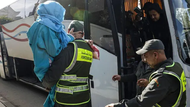 رجل أمن مغربي يحمل سيدة عجوز ترتدي ملابس مغربية زرقاء ويصعد بها على متن حافلة وخلفه رجل أمن آخر بينما يوجد ركاب في الحافلة