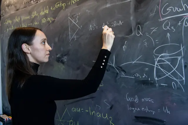 Sabrina González Pasterski con un saco negro escribe sobre un pizarrón lleno de fórmulas