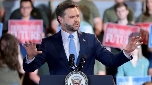 Vice-President JD Vance addresses a crowd of supporters in Maine. He is wearing a blue suit and holding his arms out, with a number of supporters sat behind him holding signs saying "Defend our Future"