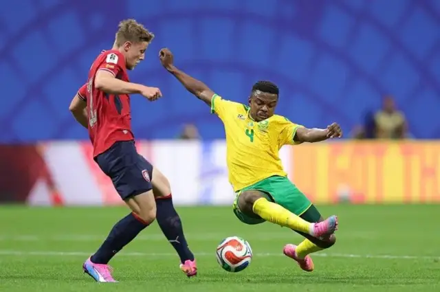 South Africa captain Tehobo Mokoena wear yellow shirt and green shorts e dey in motion wit Czech Republic player wey wear red shirt and navy blue shorts.