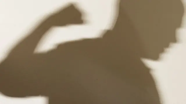 Posed image of di shadow across di wall of man wit clenched fist, wit di shadow appearing to tower ova woman wey dey cower for di corner of navy blue sofa.