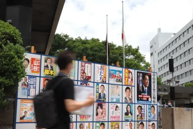 Seorang pria berjalan melewati poster kampanye kandidat Partai Demokrat Liberal (LDP) dalam pemilihan majelis tinggi, sementara di belakangnya bendera nasional Jepang (kanan) dan bendera LDP (kiri) digantung setengah tiang di markas LDP di Tokyo, Minggu (10/07).