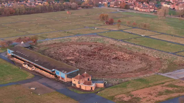 Imagen de dron de la antigua pista de carreras: un terreno de forma ovalada con una tribuna a un lado y campos verdes alrededor de la pista.