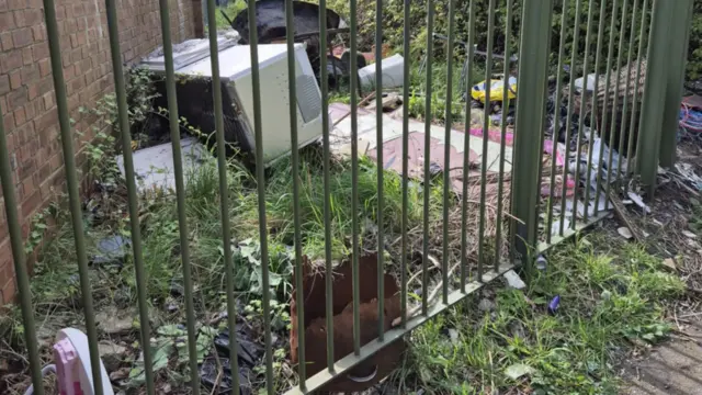 Piles of rubbish including old toys, a fridge and empty boxes thrown on a patch of grass in Grangetown. It is lying behing a green metal fence.