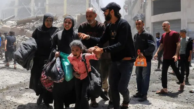 Família com pertences em meio a construções destruídas após bombardeio na Faixa de Gaza