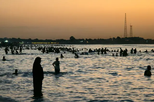 Mutane na iyo a cikin teku bayan faduwar rana. Ana iya ganin wata mata sanye da doguwar riga da hijabi tsaye cikin ruwan da ya kai har kaurin kafafunta. 