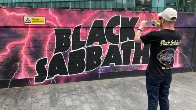 Um fã com uma camiseta do Black Sabbath tirando uma foto de um grande mural colorido do Black Sabbath em uma parede em Birmingham