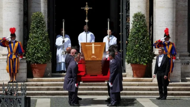 Coffin of di Pope wit a cross sign on am, wey pallbearer carry wit officials in white standing at di entrance