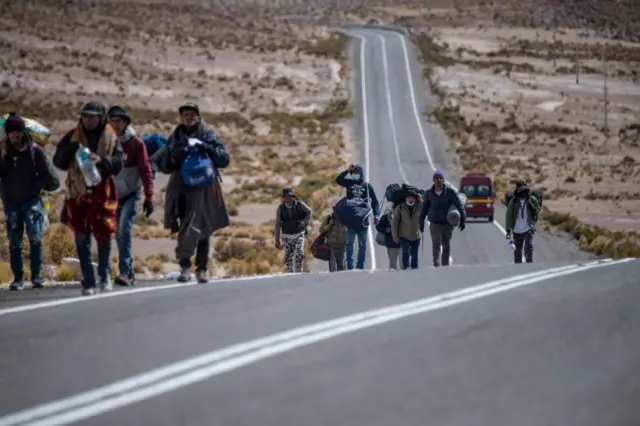Grupo de personas caminando por una carretera desértica en una zona árida, avanzando en dirección a una larga pendiente asfaltada.