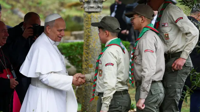 Le pape, debout à gauche, serre la main d'un scout. Deux autres personnes se tiennent à ses côtés. Ils sont à l'extérieur. Derrière le pape, un homme tient un appareil photo devant son visage.