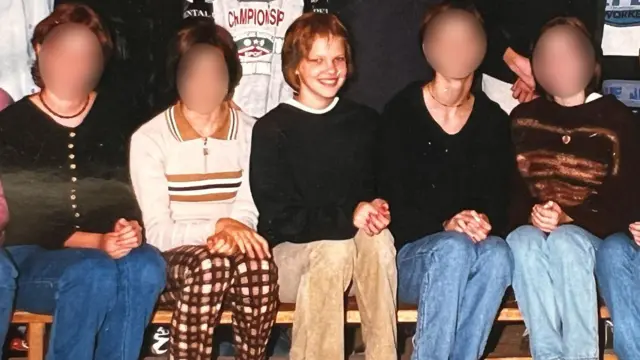 Nadia Marchinko na zdjęciu klasowym, na którym widać kilku kolegów z klasy siedzących w rzędzie na drewnianej ławce w pomieszczeniu, z rękami opartymi o kolana.
