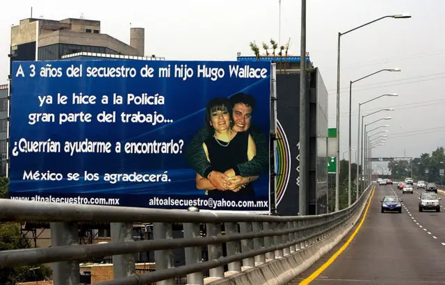 Una valla azul grande con letras blancas en el que Isabel Miranda afirma que ya ella hizo gran parte del trabajo de la policía y pide ayuda para hallar a su hijo. En la parte derecha de la valla hay una foto de Isabel Miranda siendo abrazada por la espalda por su hijo, ambos mirando hacia el frente. Al lado derecho de la valla, hay una especie de autopista en la que varias vehículos se acercan desde lejos.