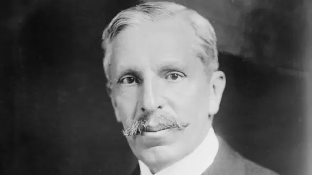 Mężczyzna z wąsami, ubrany w białą koszulę, patrzy prosto w kamerę.