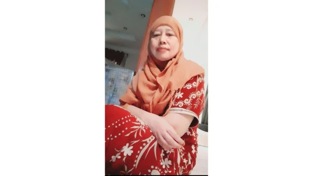 Pekerja migran Indonesia, Suniah Sahmin, duduk di depan pintu dengan daster motif warna merah bata dan kerudung cokelat.