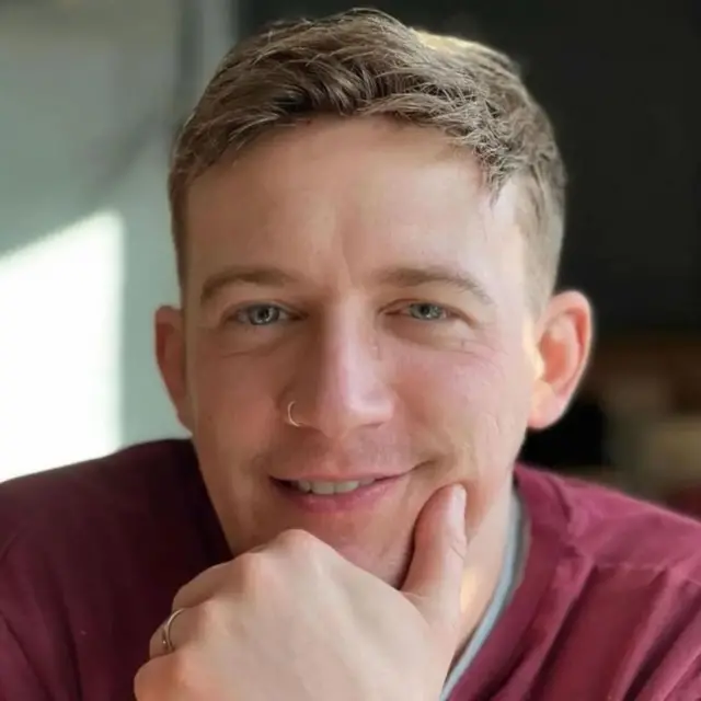 د یوه سړي څېره، چې کامرې ته ګوري، لاس یې د زنې لاندې ایښی، موسکا کوي او په پوزه کې حلقه لري.