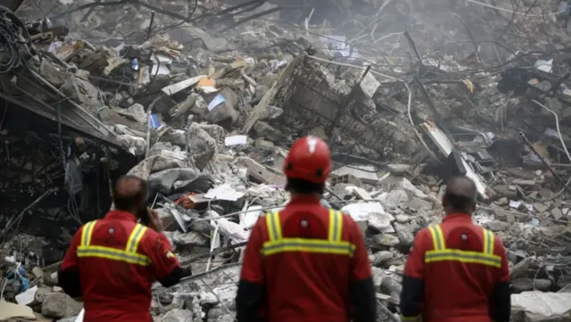 چند امدادگر پشت به دوربین و رو به ویرانه‌ها ایستاده‎اند در حال تماشای ساختمان‌های ویران شده هستند