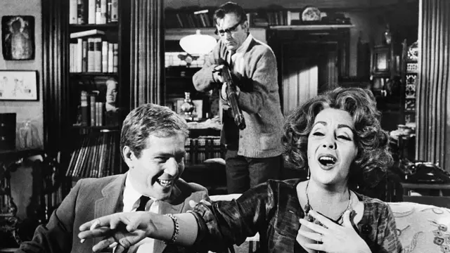 Elizabeth Taylor, Richard Burton (trzymający karabin) i George Segal na scenie z powieści Edwarda Albee'ego „Kto się boi Virginii Woolf?".