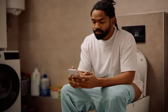Un homme noir utilisant un smartphone assis sur les toilettes.