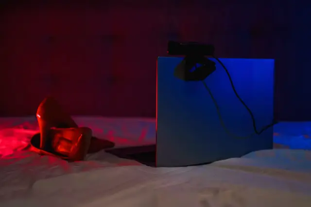 کامپیوتری با وب‌کم روی یک تخت که در کنارش یک جفت کفش پاشنه بلند زنانه دیده می‌شود