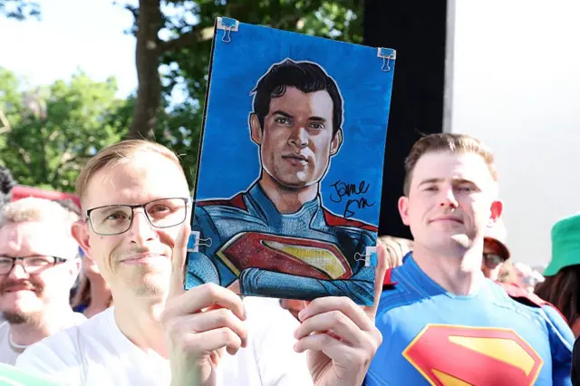Homem segurando uma foto do Superman. Ao lado dele, outro homem usa uma fantasia do Superman