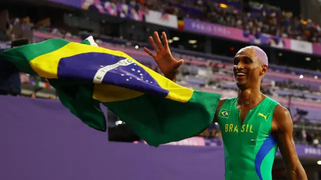 Alison do Santos, um homem pardo, ao lado da bandeira do brasil, sorrindo