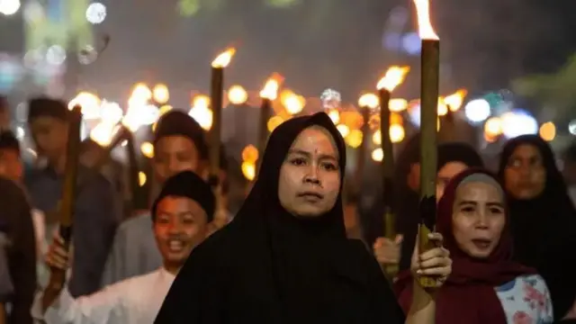 No primeiro plano, uma mulher com a cabeça coberta, com uma tocha na mão, seguida por várias pessoas atrás.