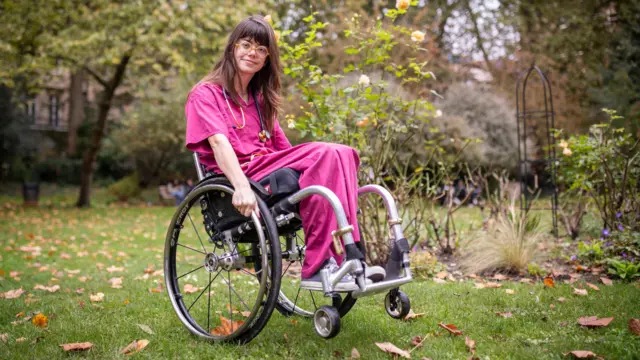 Grace Spence Green sentada em uma cadeira de rodas, vestindo um uniforme médico cor-de-rosa, em um parque