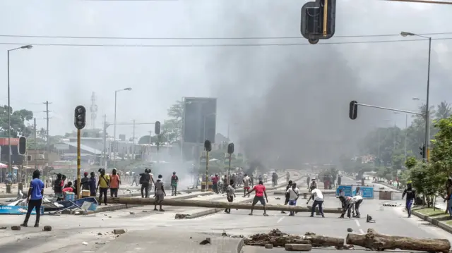 Abigaragambya bafunze imihanda bakoresheje ibiti n’ibindi bikoresho i Dar es Salaam ku wa 29 Ukwakira 2025, mu gihe habaga amatora ya Perezida wa Tanzania. Inyuma yari umwotsi urimo kuzamuka. 