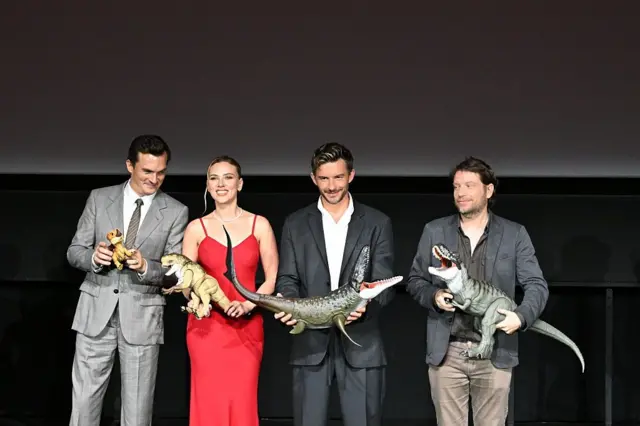Atores aparecem segurando dinossauros de brinquedo