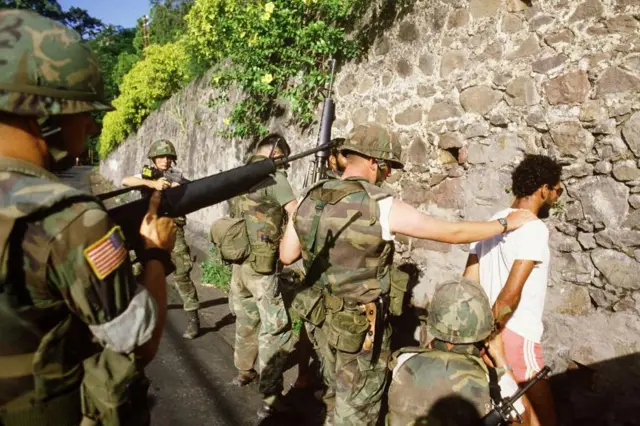 Американские солдаты обыскивают жителя Гренады, подозреваемого в причастности к марксистскому движению, 1983 год. Он стоит лицом к каменной стене, один из солдат в каске положил руку ему на плечо