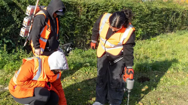 Tres trabajadores con chalecos naranjas de alta visibilidad utilizan taladros para colocar pequeños dispositivos rojos llamados nodos en el suelo herboso. Uno de ellos lleva una mochila con varios nodos adheridos. Llevan guantes y capuchas para abrigarse. Al fondo se ve un seto denso.