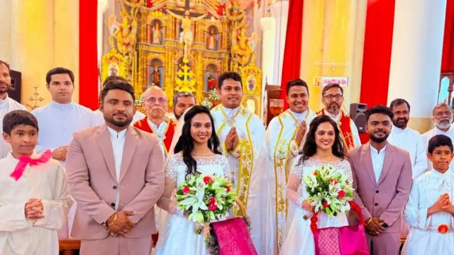 Two brides holding bouquets stand beside their newly married husbands, with a priest in a white robe between them. Twin altar boys stand at either end of the group, while relatives and guests gather behind. In the background, the church altar features a depiction of Jesus on the cross.