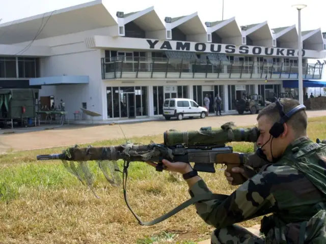 Un soldat français en position accroupie regarde dans un fusil à lunettes avec un casque à micro sur la tête. En arrière-plan un bâtiment sur lequel est écrit "Yamoussokro" devant lequel se trouvent un véhicules, des jeeps militaires et des gens.