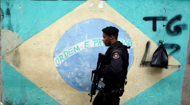 Policial passa na frente de bandeira do Brasil pintada em muro durante operação policial no Rio
