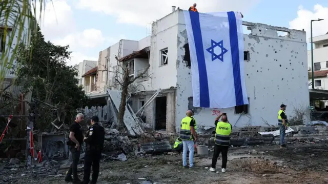 Emergency workers stand in front of a damaged building which has a large Israeli flag draped over it. විශාල ඊශ්රායල ධජයක් අතුරා ඇති හානියට පත් ගොඩනැගිල්ලක් ඉදිරිපිට හදිසි සේවකයින් සිටගෙන සිටියි.