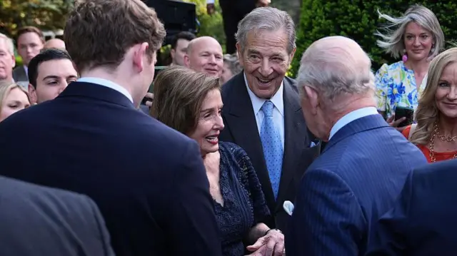 Nancy Pelosi in a blue dress chats with the King in a pinstripe suit at the garden party in the gardens of the British Embassy in Washington. Other guests watch on in the background. 