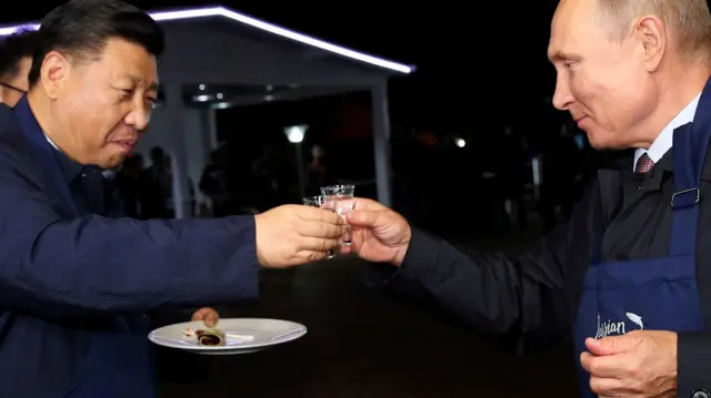 Çin Devlet Başkanı Şi Cinping elinde bir tabak tutuyor, Rusya Devlet Başkanı Vladimir Putin ise üzerinde bir önlükle ve iki lider ellerinde küçük bardak tokuşturuyor 