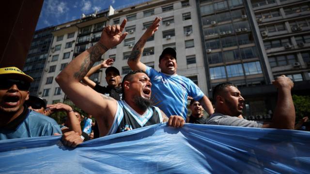Homens erguendo os braços na rua, usando camisas e bandeira da Argentinaprotesta contra las medidas del presidente Javier Milei