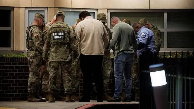 شماری از سربازان گارد ملی امریکا به مقابل شفاخانه‌ای که سربازان زخمی بستر هستند برای دعا گردهم آمده اند. سربازان برخی شان یونیفرم نظامی بر تن دارند. سرشان به علامت دعا پایین است.