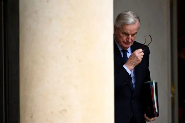 Michel Barnier, bir kolonun arkasında, başını öne eğmiş, gözlüğünü çıkararak yürüyor.