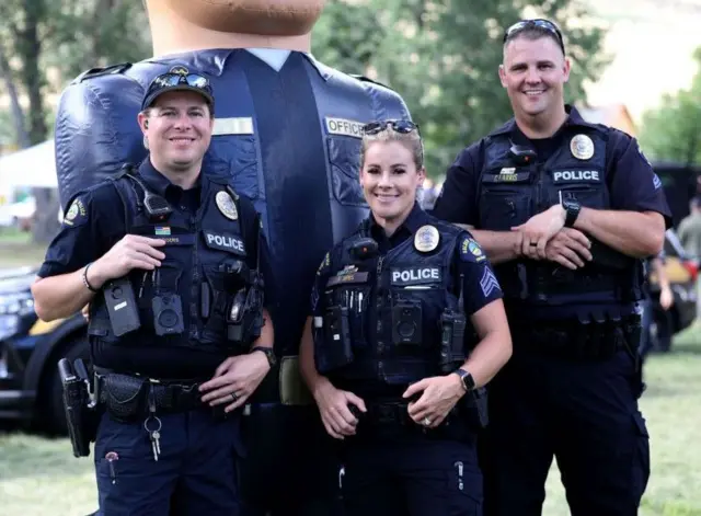 دوه نارینه افسران او یوه ښځینه افسره، چې یونیفورم یې په تن دی د کولوراډو په ګولډن ښار کې په یوه سیمه ییز پارک کې ځان برابر کړی، چې انځور یې واخیستلی شي.