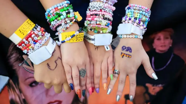 A imagem mostra quatro pessoas lado a lado exibindo os pulsos repletos de pulseiras coloridas de miçangas e ingressos de um show. Os acessórios, muitos deles com palavras e referências a músicas, aparecem ao lado de unhas coloridas, anéis e pequenas pinturas nas mãos, destacando uma tradição entre os fãs da cantora de confeccionar e trocar pulseiras durante os eventos.