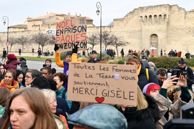 Pessoas seguram cartazes em frente ao tribunal de Avignon, na França, em 19 de dezembro de 2024, com o dizeres: "'Natal na prisão, Páscoa na prisão' e 'Todas as mulheres do mundo apoiam você, obrigada, Gisèle'