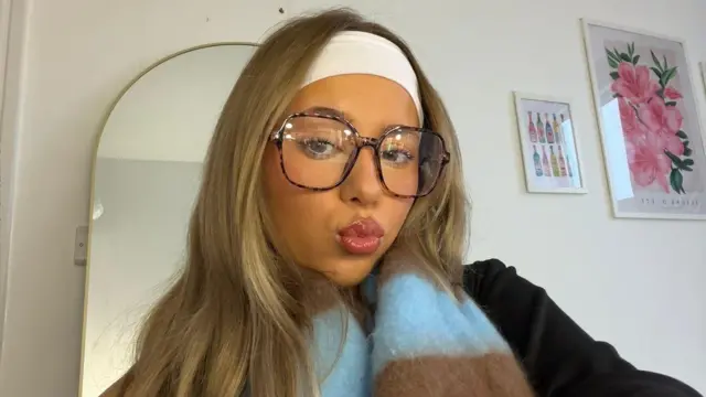 Chloe, z długimi prostymi blond włosami, robi dzióbek do selfie, a za nią widać lustro.