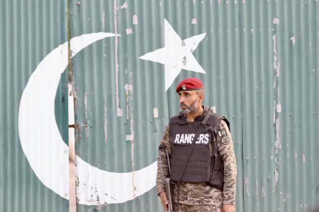 یک نظامی مسلح پاکستان کنار دیواری که با پرچم رنگ‌آمیزی شده است.