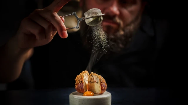 Un hombre espolvorea condimentos sobre un plato de comida muy bien presentado. Sostiene la mano sobre el plato y el condimento cae sobre él. El rostro de un hombre se ve en el fondo oscuro.