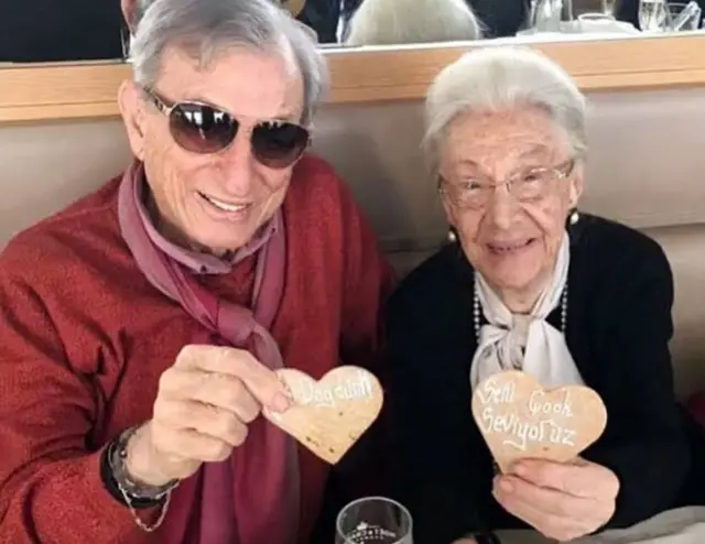 Yaş almış bir kadın ve erkek ellerine kalp şeklinde kurabilerle kameraya gülümsüyor.